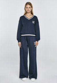 Felpa blu navy con strisce bianche sul collo a V e sul bordo, abbinata a pantaloni a gamba larga coordinati. Tessuto morbido con dettaglio del logo.