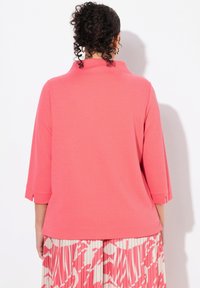 Maglietta lunga a maniche con un vivace colore corallo e un motivo testurizzato, presenta un colletto alto e spacchi laterali, abbinata a una gonna plissettata nei toni del rosa e crema.