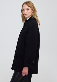 Cappotto di lana nero con colletto, tasche frontali e dettagli in bottone. Il design è oversize con una texture liscia e una silhouette sartoriale.
