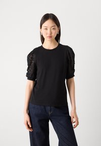 Μαύρη μπλούζα με διάφανες φουσκωτές δαντελένιες μανίκες; συμπαγές σώμα, στρογγυλή λαιμόκοψη και ελαφρώς χαλαρή εφαρμογή. Συνδυάζεται με σκούρα μπλε τζιν παντελόνια.