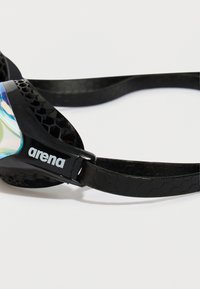 Detailný záber na čierny remienok plaveckých okuliarov s hexagonálnym vzorom a logom "arena" po boku, na bielom pozadí.