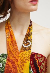 Femme portant une robe halter multicolore avec des motifs complexes orange, jaune, rouge et vert, montrée du cou à la partie supérieure de la poitrine sur un fond blanc.