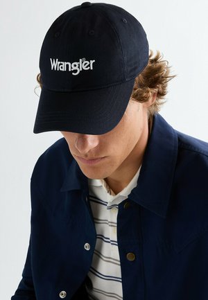 Młody mężczyzna o kręconych włosach, noszący ciemnogranatową czapkę Wrangler, granatową kurtkę i białą koszulkę polo w paski, patrzący w dół.