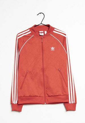 Czerwona kurtka Adidas zapinana na zamek błyskawiczny z białymi paskami na rękawach, dwoma kieszeniami na zamek i logo na górnej lewej stronie klatki piersiowej, wisząca na czarnym wieszaku.