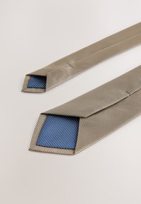 Texturierte Krawatte in Beige mit einem blauen gepunkteten Futter. Verfügt über ein schmales, spitzes Ende und eine glatte Oberfläche. Geeignet für formelle Kleidung.