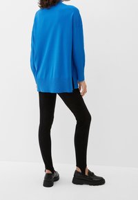 Portant un pull ample bleu vif, des leggings noirs ajustés avec des fentes aux chevilles, et des mocassins noirs, debout contre un mur blanc.