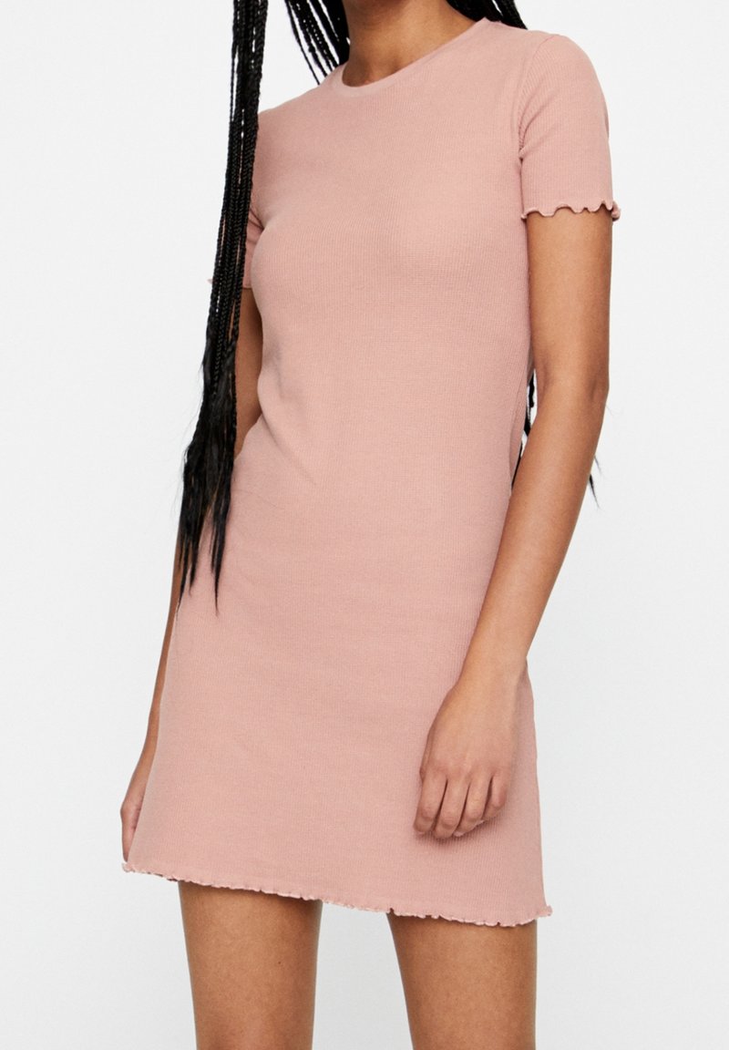 Femme portant une robe courte à manches courtes, côtelée, de couleur rose clair avec des bords festonnés aux manches et à l'ourlet, sur un fond blanc uni.