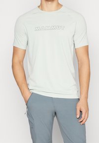 Jasnoszara koszulka z krótkim rękawem, wykonana z gładkiego, elastycznego materiału, z okrągłym dekoltem i logo "MAMMUT" w kontrastowym kolorze z przodu.