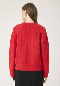Woman with straight brown hair wears bright red cable-knit sweater and black skirt, facing away against light background.