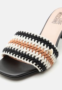 Μαύρο σανδάλι mule ανοιχτού δακτύλου με φαρδιά πολύχρωμη λουρίδα πλεκτή σε μαύρο, λευκό και ταμπά, με μικρό χρυσό διακοσμητικό λογότυπο σε σχήμα καρδιάς.