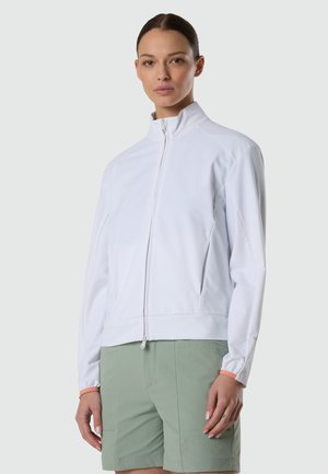Donna con giacca bianca con zip e pantaloncini verde chiaro, in piedi davanti a uno sfondo grigio uniforme.