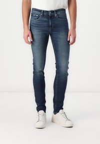 Jeans de mezclilla oscuros, de corte ajustado y con un ligero desgaste, diseño de cinco bolsillos, remaches de metal y cierre con cremallera, combinados con zapatillas blancas.