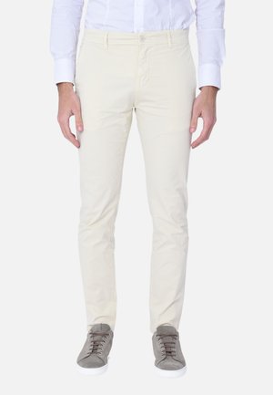 Uomo che indossa pantaloni beige aderenti, camicia bianca e sneakers grigie, in piedi davanti a uno sfondo bianco semplice.