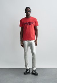 Tricou roșu din bumbac cu textul negru "DSQUARED2 MILANO", asortat cu blugi argintii strălucitori și adidași negri cu dungi albe.