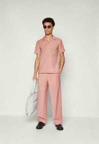 Uomo che indossa una camicia a maniche corte rosa con bottoni e pantaloni larghi abbinati, tiene in mano una borsa tote bianca imbottita, indossa occhiali da sole e scarpe nere.