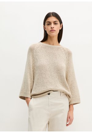 Kobieta z długimi ciemnymi włosami, ubrana w beżowy sweter o luźnym splocie i jasne spodnie, stojąca z jedną ręką w kieszeni na białym tle.