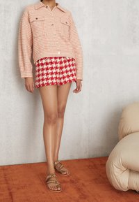 Růžový houndstooth svetr s předními kapsami, spárovaný s červenými a bílými houndstooth šortkami. Hnědé sandály s kulatými akcenty. Hladká textura.