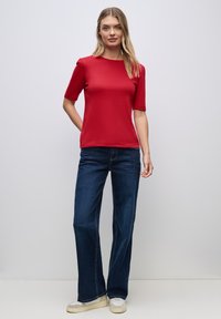 Rød kortærmet top med rund halsudskæring, parret med mørkeblå flagrende jeans og beige sneakers. Glat tekstur, minimale designdetaljer.