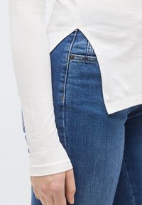 Fehér, hosszú ujjú ing oldalsó sliccel, kék farmernadrággal párosítva. Simogatós anyagtextúra; az ing testhez simuló szabású.