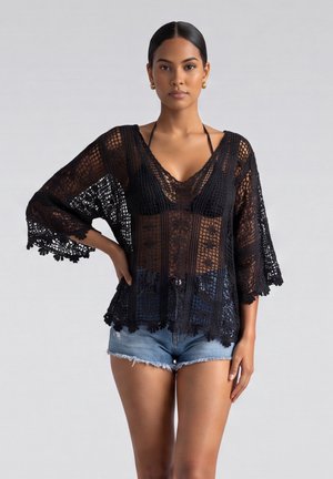 Jonge vrouw draagt een doorschijnende zwarte gehaakte top over een zwart bikinibovenstuk, gecombineerd met blauwe denimshorts, staande met één hand op de heup.