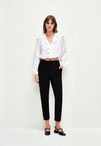 Kvinna klädd i en vit volangblus och svarta byxor i ankellängd med svarta låga klackskor, stående mot en enfärgad vit bakgrund.