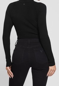 Schwarzer gerippter Rollkragenpullover mit langen Ärmeln, kombiniert mit schwarzen hochgeschnittenen Jeans, die zwei Gesäßtaschen und dezente Markenkennzeichnung aufweisen.