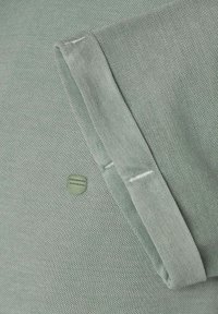 Green textured fabric with a folded sleeve, featuring a circular accent. Stitching detail visible near the fold. Casual design.