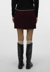 Jupe plissée bordeaux courte avec une texture côtelée, associée à des bottes en cuir noir hauteur genoux et des chaussettes beiges, vue de dos.
