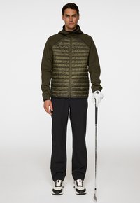 Man som bär en olivgrön fodrad jacka, svarta byxor, en vit golfhandske, svartvita skor, och håller en golfklubba mot en enkel bakgrund.