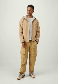Giacca leggera beige con cappuccio, abbinata a pantaloni cargo beige con diverse tasche e scarpe sportive grigie con lacci a fantasia.