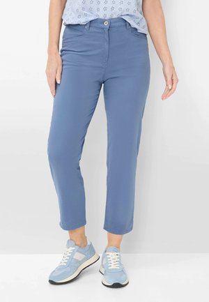 Kvinde iført korte blå bukser, lyseblå mønstret bluse og lyseblå og hvide sneakers, stående mod en hvid baggrund.