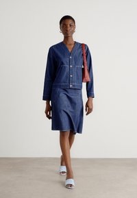 Denimjakke med knappelukking og to lommer, sammen med et matchende denimskjørt. Tilbehør inkludert en rød håndveske og blå sandaler.