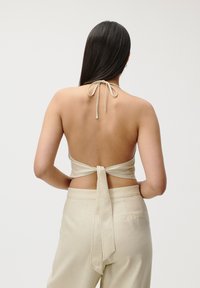 Vue arrière d'un top halter en tissu beige, avec une fermeture à nouer au cou et un design dos nu, associé à un pantalon beige.