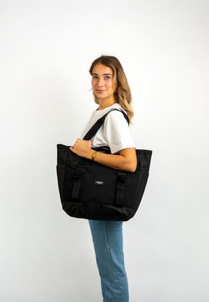 Black tote bag with two wide straps, textured fabric, and two side pockets. Features brand logo on the front and a solid color design.