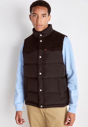 Black puffer vest with a corduroy collar, snap buttons, and front pockets. Worn over a light blue long-sleeve shirt.