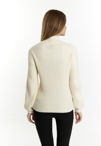 Crème ribgebreide trui met een hoge kraag, verlaagde schoudernaden en aansluitende manchetten. Gecombineerd met zwarte broek, gezien vanuit de achterkant.