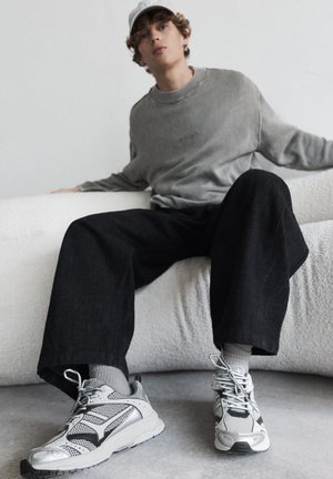 Young person in gray sweater, wide black pants, white sneakers, and cap sits on white curved sofa against plain wall.