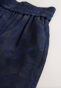 Tissu bleu marine avec un motif floral subtil, doté d'une taille élastique et d'une texture lisse, présentant des plis drapés et des détails en surface.