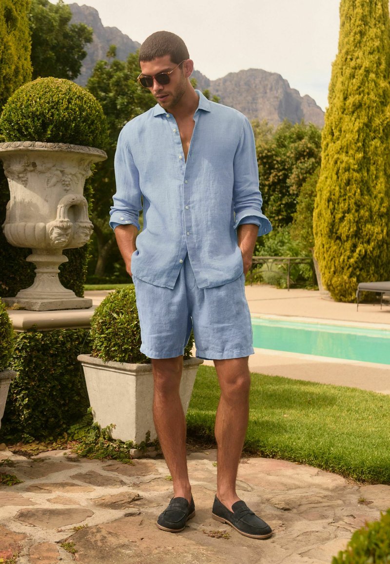 Homme portant une chemise en lin bleu clair et un short, debout près de la piscine dans un jardin luxuriant avec des montagnes en arrière-plan, mains dans les poches, portant des lunettes de soleil.