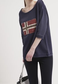 Felpa a maniche lunghe blu navy con un design testurizzato della bandiera norvegese in rosso e oro sul davanti. Indossata con pantaloni scuri, tiene una borsa grigia.