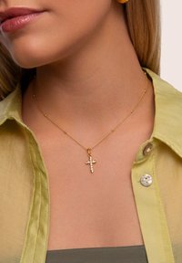 Collana d'oro con pendente a croce, caratterizzata da pietre preziose trasparenti e dettagli arrotondati. Indossata sopra una camicia verde chiaro.
