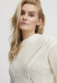 Cream knit sweater featuring a textured pattern, high neckline, and decorative detail along the shoulders, creating a cozy appearance.