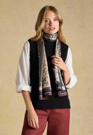 Blond kvinde iført en sort ærmeløs sweater over en hvid bluse, et mønstret tørklæde om halsen og røde bukser, stående indendørs.