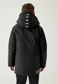 Parka noire avec capuche, arborant une étiquette "BAKER" sur le col, en matériau texturé, et un patch logo près de l'ourlet.