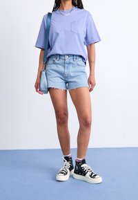 Maglietta viola a maniche corte con tasca, pantaloncini di jeans azzurri con orlo sfrangiato e sneaker alte nere con suole bianche.