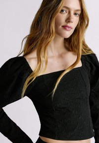 Top nero strutturato con scollo quadrato e maniche a palloncino. Caratterizzato da un design aderente e un sottile motivo floreale su tutto il tessuto.