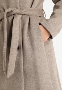 Beige wool coat featuring a textured, herringbone pattern, front buttons, and a tie waist with side pockets.