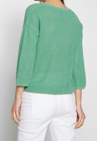 Femme portant un pull en tricot vert à manches trois-quarts et un pantalon blanc, vue de dos contre un fond uni.