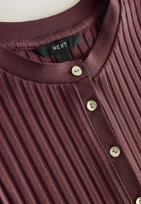 Top burdeos acanalado con cuello redondo, con tres botones de tono dorado y una etiqueta con la palabra "NEXT" sobre un fondo negro.