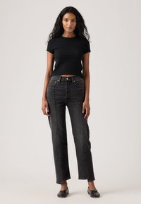 Kvinde med langt krøllet hår iført en sort top med korte ærmer, mørke højtaljede, lige jeans og sorte flade sko stående foran en neutral baggrund.